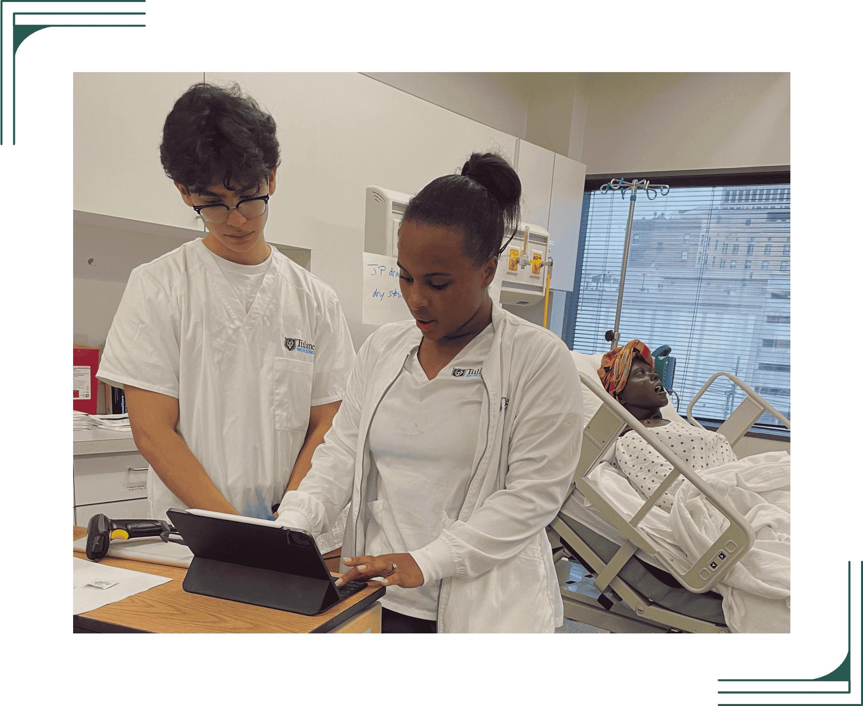 Two nursing students use a tablet next to a hospital bed with a patient mannequin.