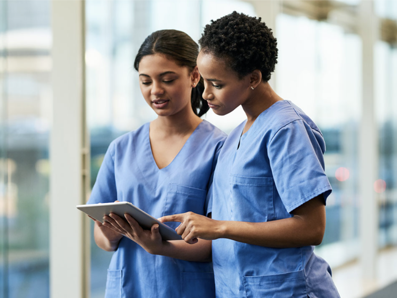 Two nurses in blue scrubs focused on a tablet, one pointing at the screen.