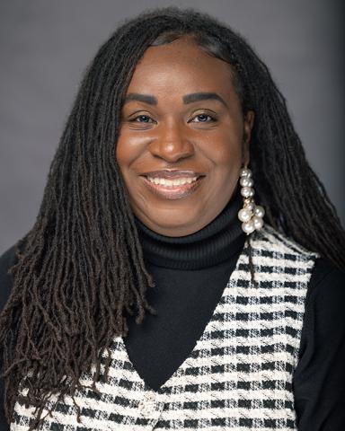 Smiling woman with long dreadlocks, black turtleneck, and black and white plaid vest.