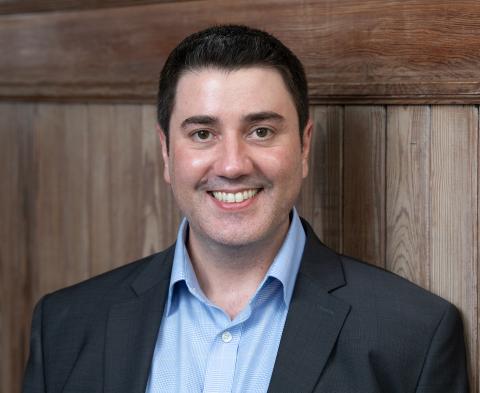 Smiling man in blue shirt and dark suit jacket against a wall