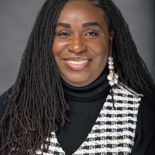 Smiling woman with long dreadlocks, black turtleneck, and black and white plaid vest.
