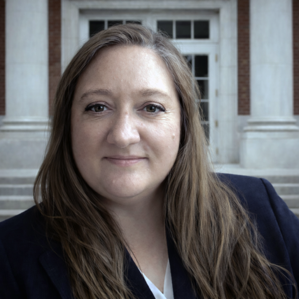 Woman with long brown hair, pleasant expression, classical building background.