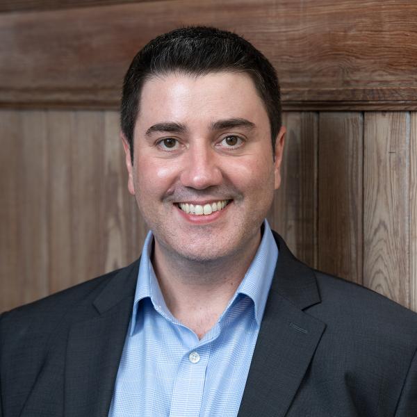 Smiling man in blue shirt and dark suit jacket against a wall
