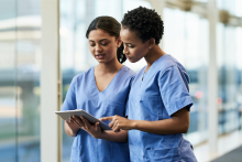 Two nurses in blue scrubs focused on a tablet, one pointing at the screen.